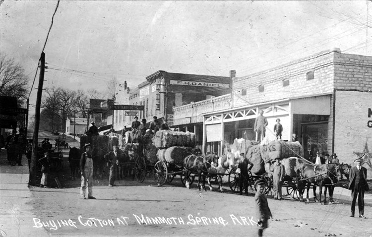 Men transporting cotton bales with horse-drawn wagons on town street with multistory storefronts