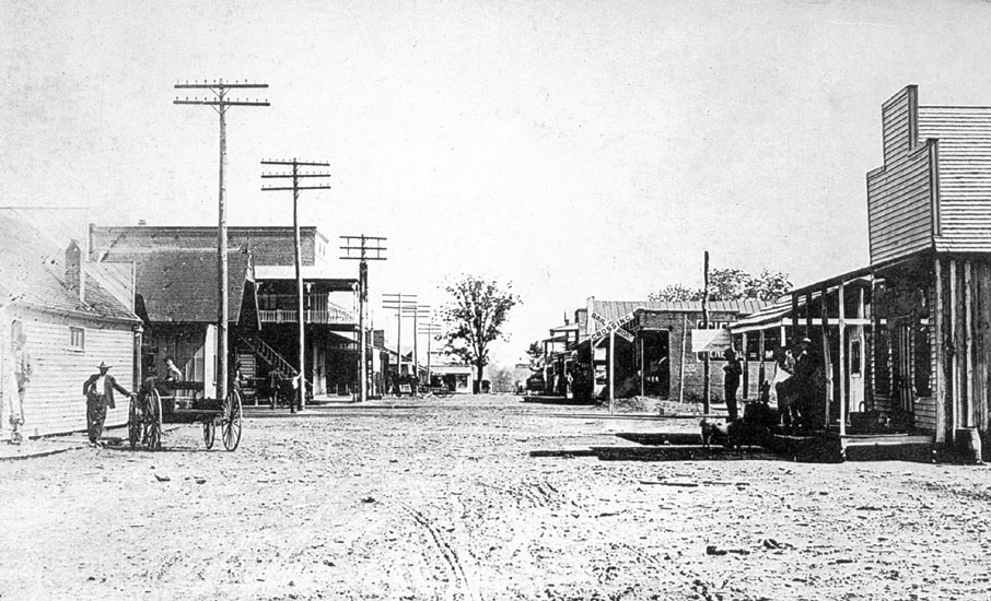 Men pose with wagon on dirt road lined with buildings and telephone poles