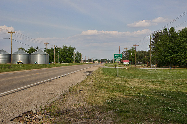 Three silos and power lines on two-lane road across from "Manilla" road sign