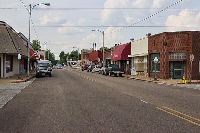 Two-lane road with single-story storefronts and parked cars on both sides