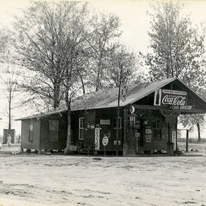 Single-story service station building with covered entrance and two gas pumps on dirt road