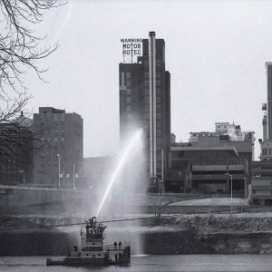 Multistory skyscrapers and other buildings as seen from across river with boat spraying water toward the "Manning Motor Hotel"