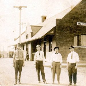 Four white men standing in line with building with "Mansfield" sign behind them