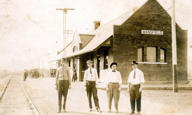 Four white men standing in line with building with "Mansfield" sign behind them