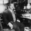 African-American man with mustache in suit vest and tie sitting at desk with book