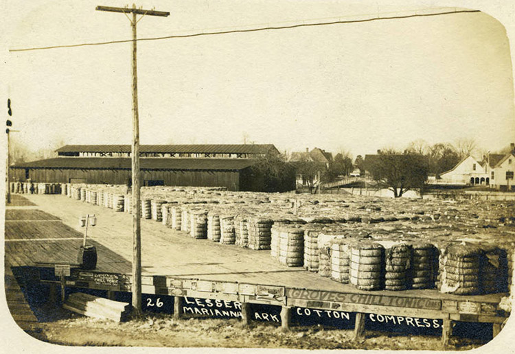 Stacks of cotton bales sitting outside warehouse building with houses in the background