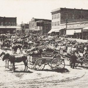 Group of horse drawn wagons on street with multistory buildings in the background