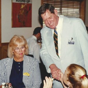 White man in suit standing shaking hands with older white woman and young white girl at table