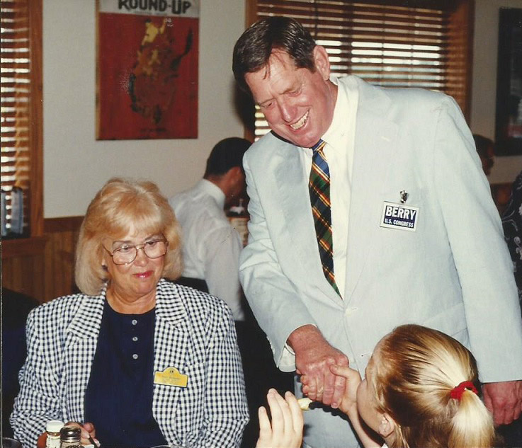 White man in suit standing shaking hands with older white woman and young white girl at table
