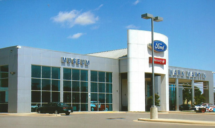 Race cars parked outside single-story museum building with Ford logo covered entrance and large windows on parking lot