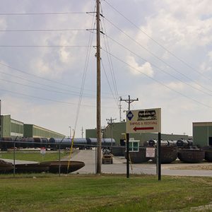 Green and white industrial buildings and train inside fence with power lines and road sign