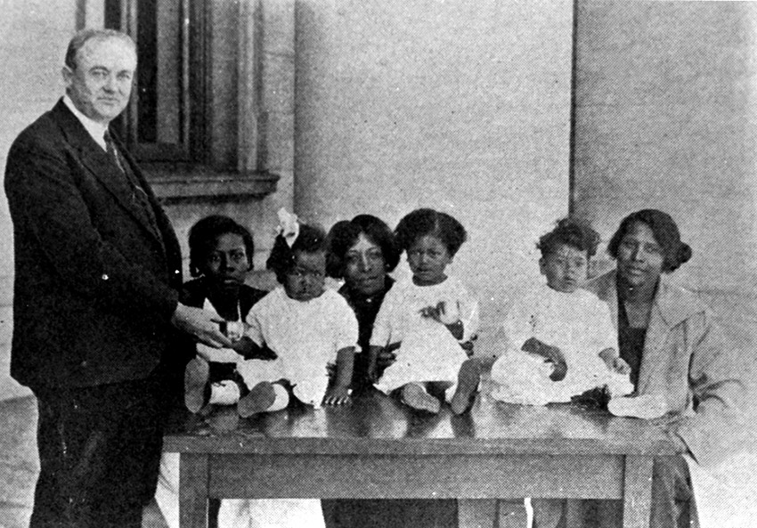 White man standing in suit with African-American women and children sitting beside him