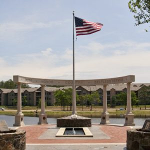 Columns and rounded top with the words "duty honor country selfless service" and flag pole with lake and apartment buildings in the background