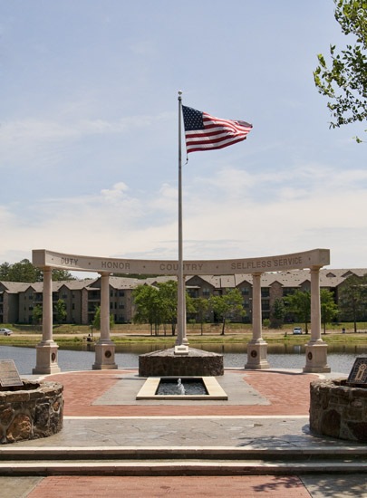 Columns and rounded top with the words "duty honor country selfless service" and flag pole with lake and apartment buildings in the background