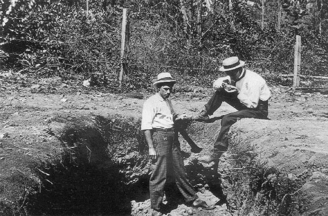 White man in hat standing in pit with another man in a hat sitting on the ground with fence behind them