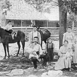 White men woman and children standing under tree with horses outside log cabin with covered porch