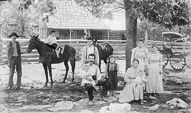 White men woman and children standing under tree with horses outside log cabin with covered porch