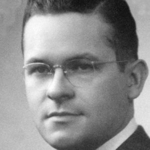 Younger white man with glasses in suit and tie and combed back dark hair