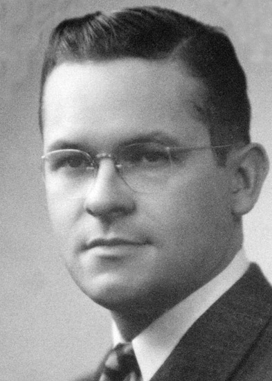 Younger white man with glasses in suit and tie and combed back dark hair