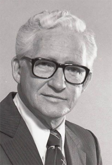 White man with white hair and black-rimmed glasses in suit and tie
