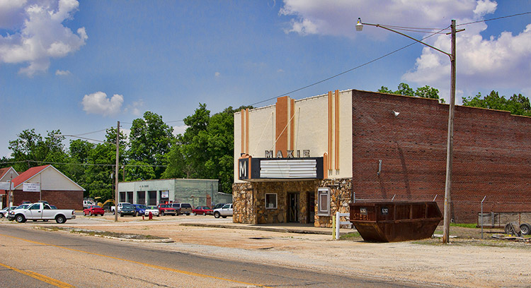 Brick and stone "Makie theater" building on multilane street with brick buildings and garage building in the background