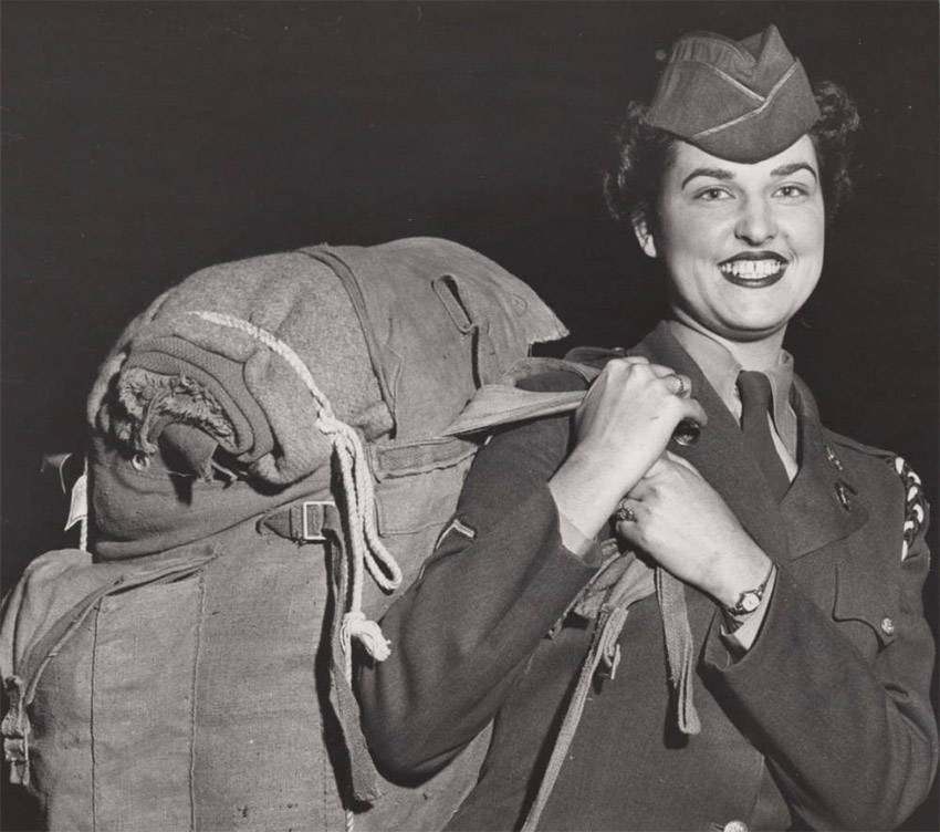 White woman wearing military uniform holding large back by straps over right shoulder