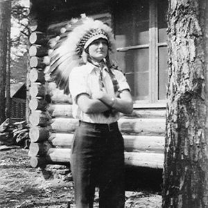 White woman wearing Native American headdress with arms crossed standing outside log cabin