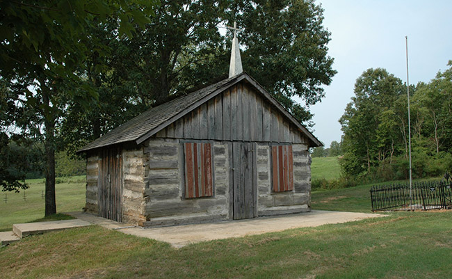 small log cabin church with steeple and cross on grass with trees