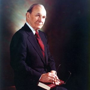 Old white man in pinstriped suit holding a pair of glasses