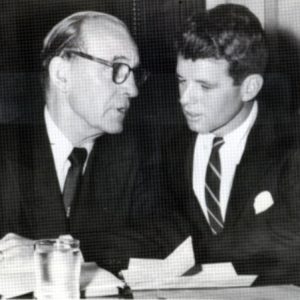 Older white man in glasses and suit speaking with young white man in suit at table