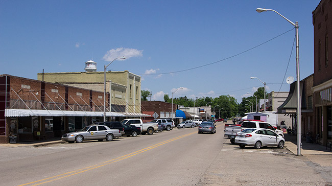 Street with brick storefronts parked cars and water tower in the background