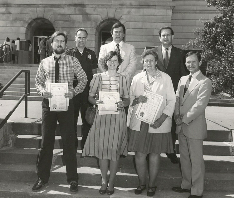 White policeman and three white men in suits posing with white man and women with papers in their hands