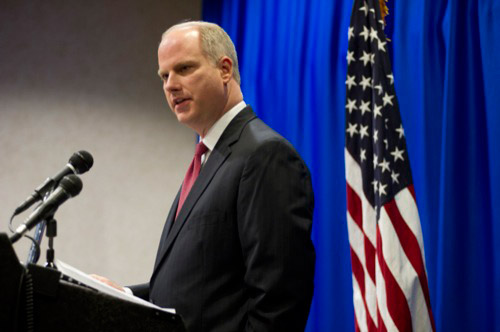 White man in suit speaking at lectern with microphone with American flag behind him and blue curtain