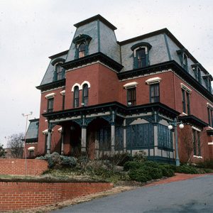 Three-story ornate brick house with central tower section and covered porch with brick walls in front and steep driveway