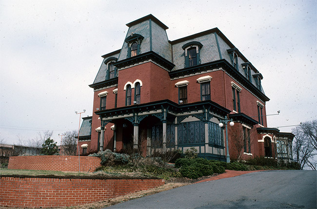 Three-story ornate brick house with central tower section and covered porch with brick walls in front and steep driveway