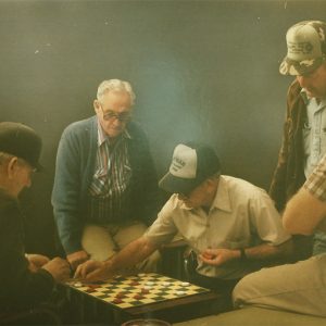 Group of older white men watching two men play checkers