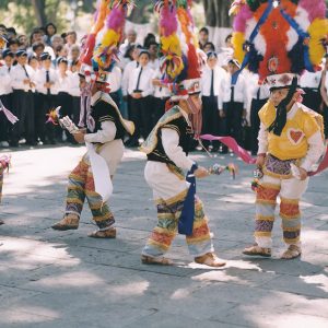 Mexican children dancing in traditional garb with crowd of children in white shirts and dark ties visible behind them