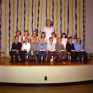 White woman in white dress on stage with class of white children in formal attire seated in front of her