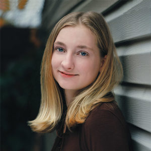 Young white girl with shoulder-length blond hair smiling while leaning against a wall