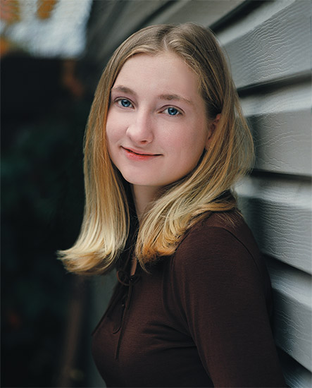 Young white girl with shoulder-length blond hair smiling while leaning against a wall