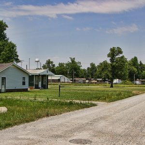 Single-story houses on gravel road with trees and water tower in the background
