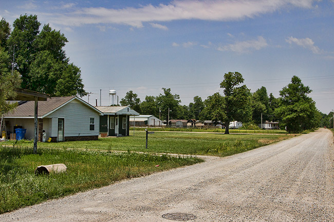 Single-story houses on gravel road with trees and water tower in the background