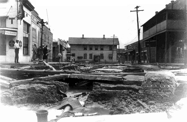 Damaged street surrounded by two-story town buildings with people on sidewalk