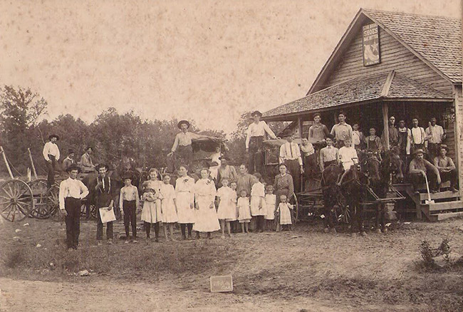Crowd of white men women and children outside single-story building with covered porch