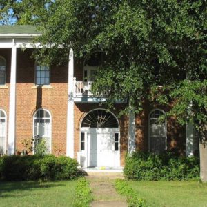 Two story brick  home with columns, balcony, arched windows and door, viewed from front yard with tree