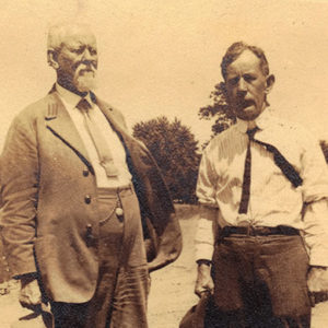 Old white man in suit with hat in right hand standing next to younger man in shirt and tie with hat in right hand