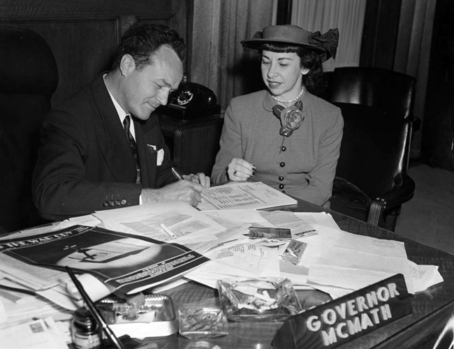White man in suit signing paper on cluttered desk for white woman in hat and pant suit in his office