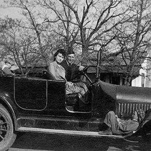 White man and woman in convertible car with tree and house in the background