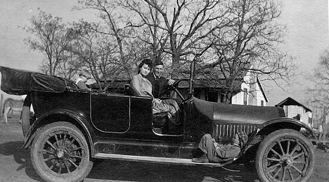 White man and woman in convertible car with tree and house in the background