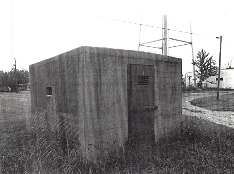 Small concrete building with iron door and water tower in the background